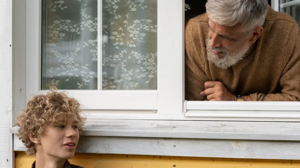 Elderly man with white beard interacts with curly-haired teenager through open window.
